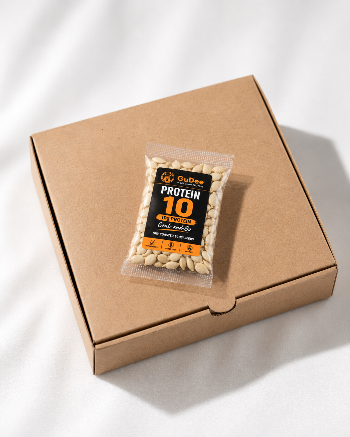 A clear bag of GuDee Protein 10 gran-and-go protein pack sits on top of a brown kraft paper box against a white background with soft shadow patterns.